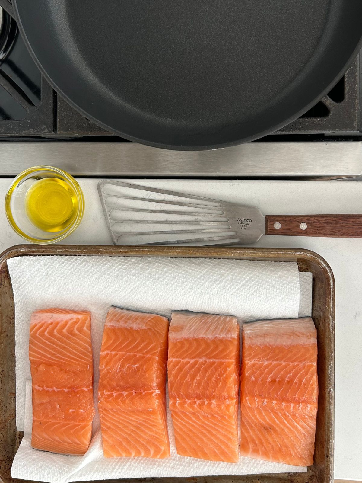 Brined, rinsed, and dried salmon fillets on a towel-lined rimmed baking sheet, oil, and a spatula next to a skillet heating up over a burner.