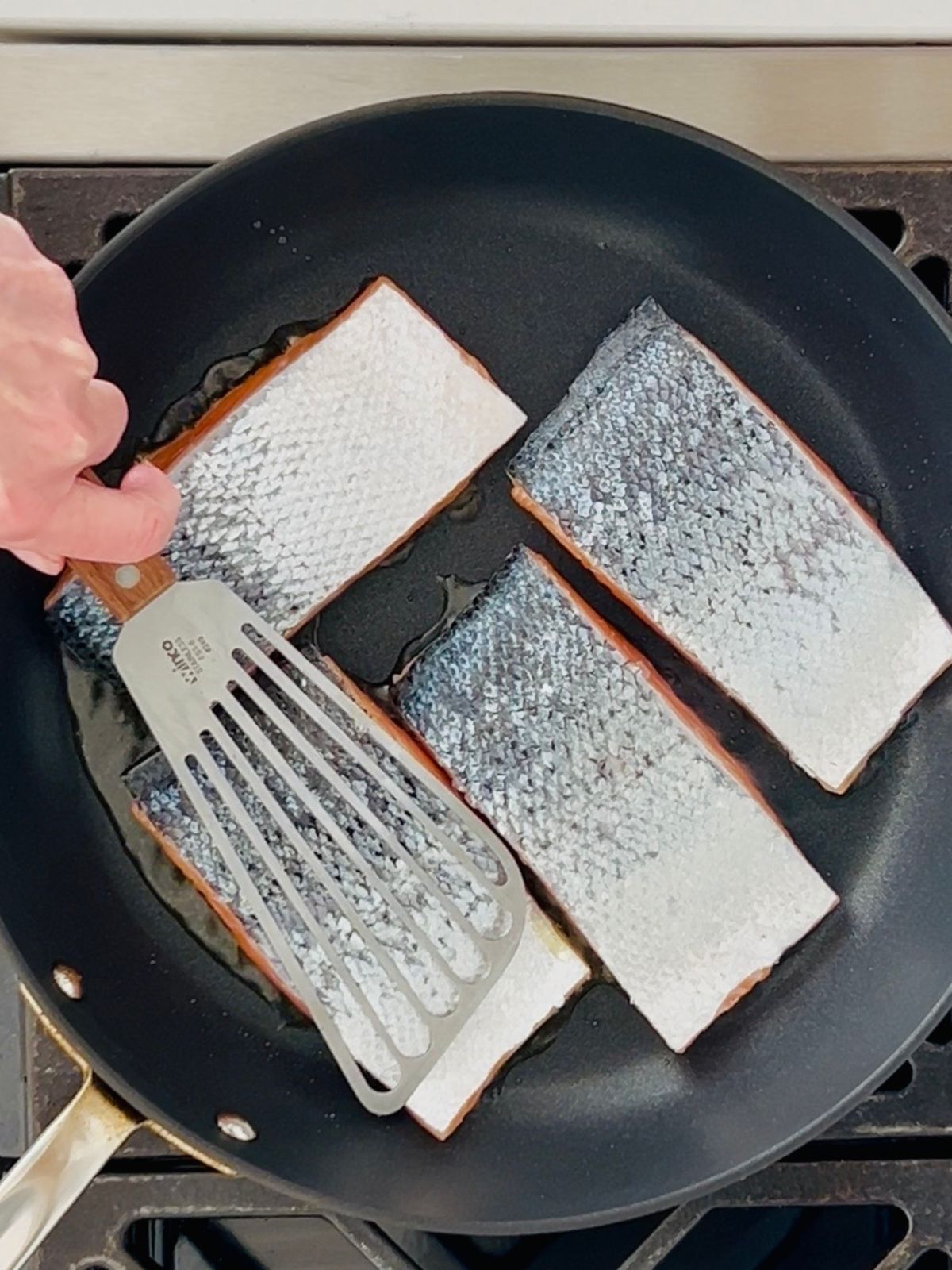 A spatula is pressing down on the skin-side of salmon fillets while they are cooking in a skillet.