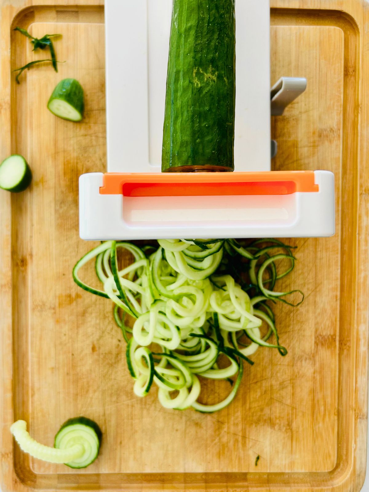 Cucumber noodles coming out of a countertop spiralizer onto a bamboo cutting board.