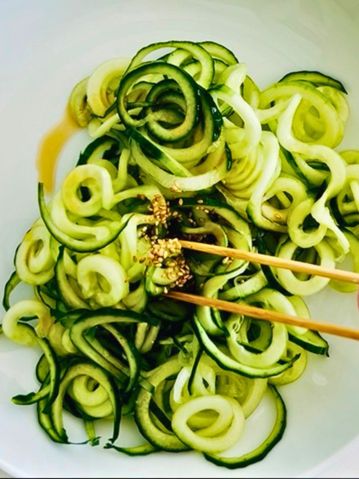 A white bowl of spiralized cucumber noodles topped with sesame seeds and tossed with sesame oil.