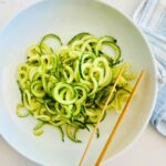 Spiralized cucumber noodles in a white bowl with chop sticks resting on the side.