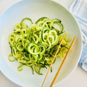 Spiralized cucumber noodles in a white bowl with chop sticks resting on the side.
