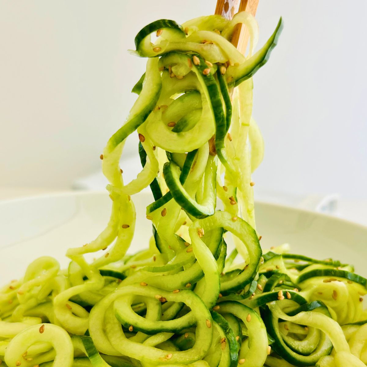 Wooden tongs lifting up a serving of cucumber noodles from a white bowl.