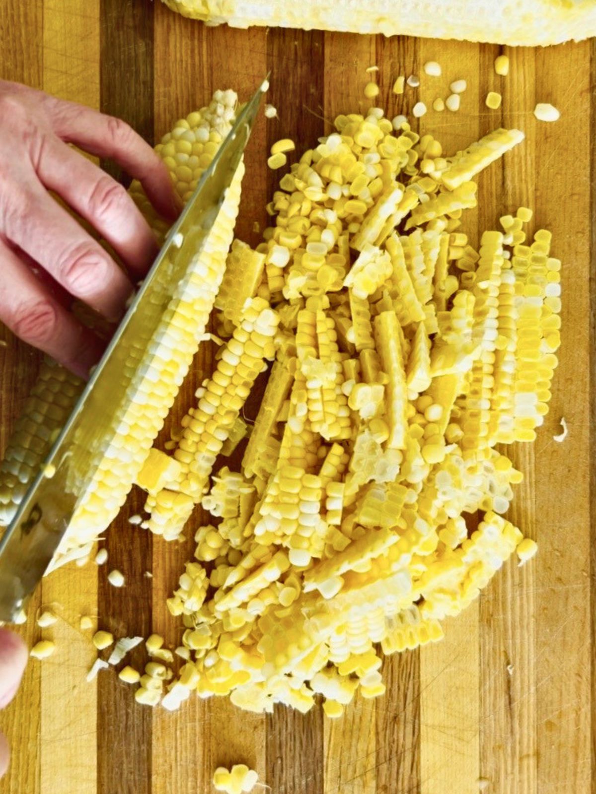Cutting fresh corn kernels off the side of a cob on a wood cutting board to prepare for corn cob stock.