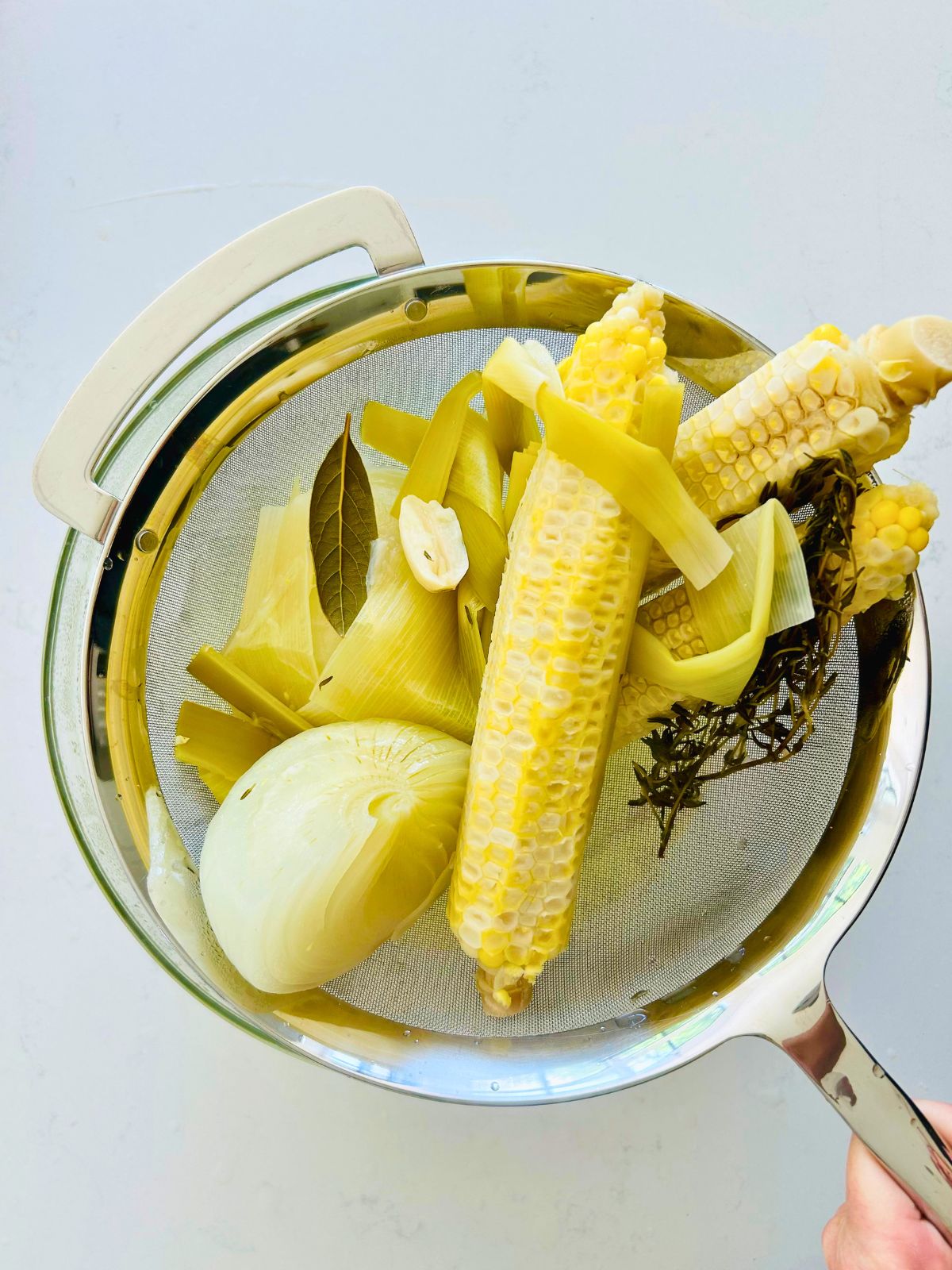 Straining spent corn cobs, onion, leeks, garlic, and herbs from finished corn broth into a glass bowl.