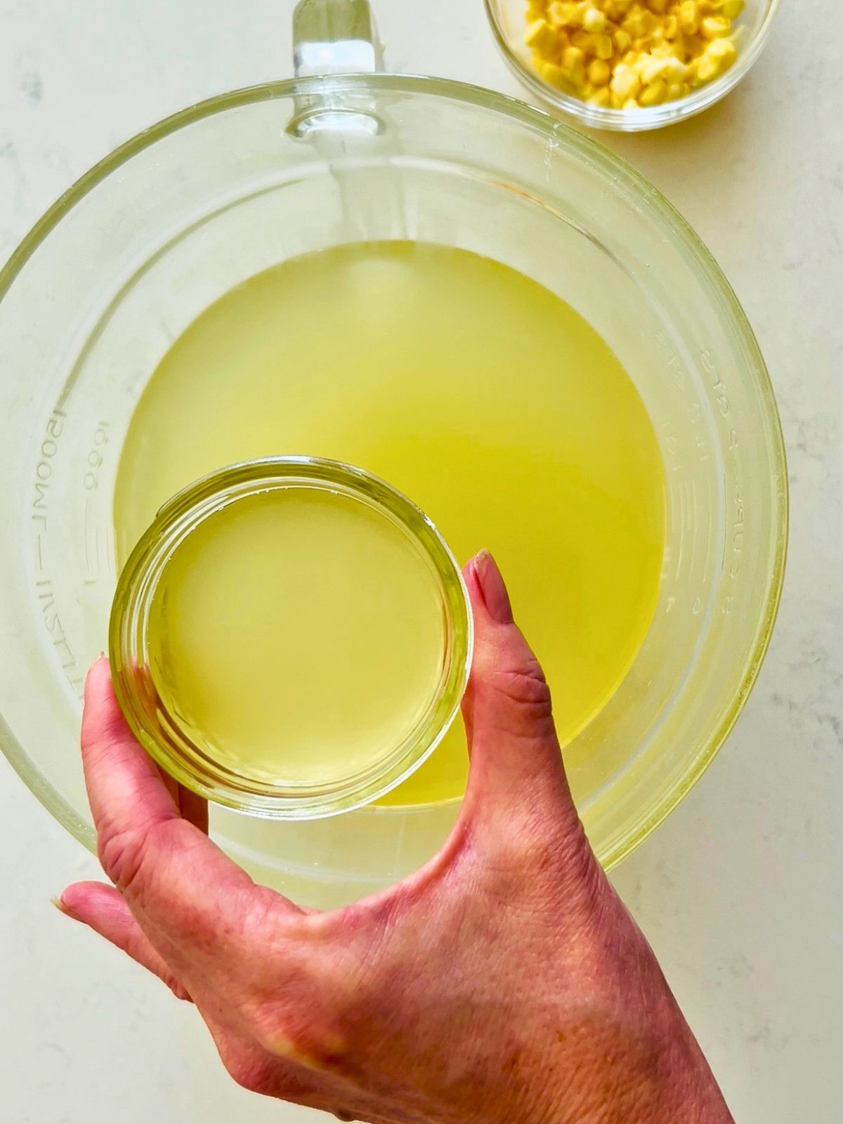 A hand holds a small mason jar of golden corn stock recipe liquid next to a dish of fresh corn kernels