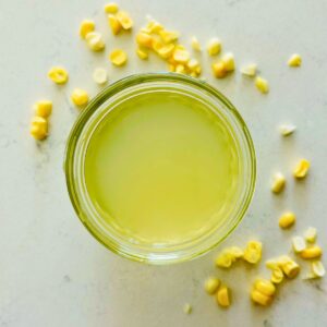 A small mason jar with corn stock next to corn kernels on a white marble surface.