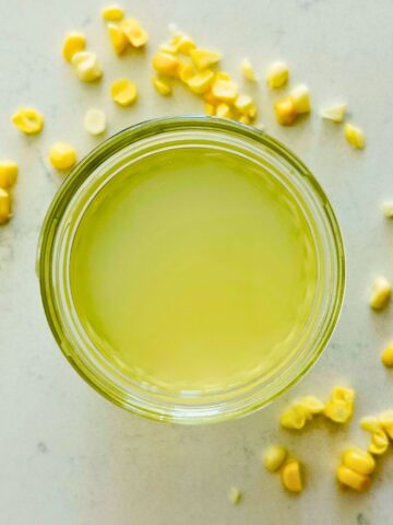 A small mason jar with corn stock next to corn kernels on a white marble surface.
