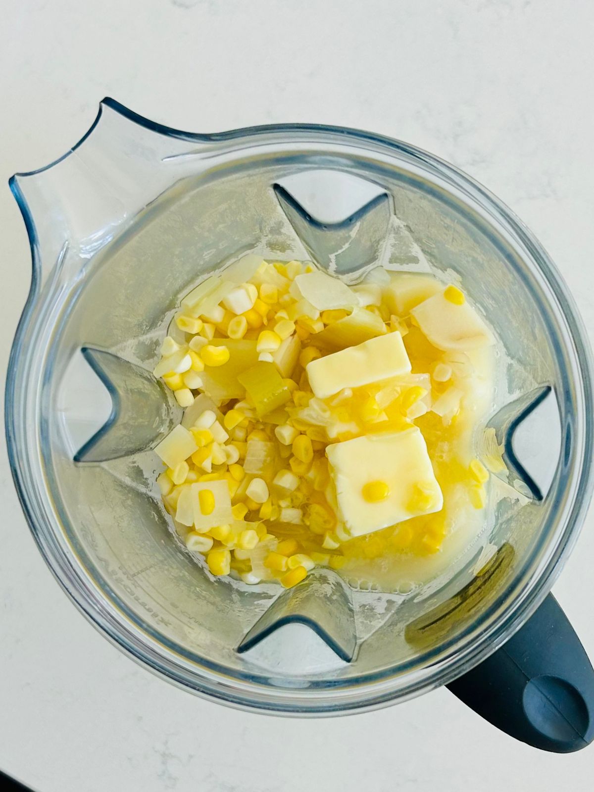 Cooked corn, potatoes, onions, leeks, and butter are in the blender's pitcher to puree into corn soup.