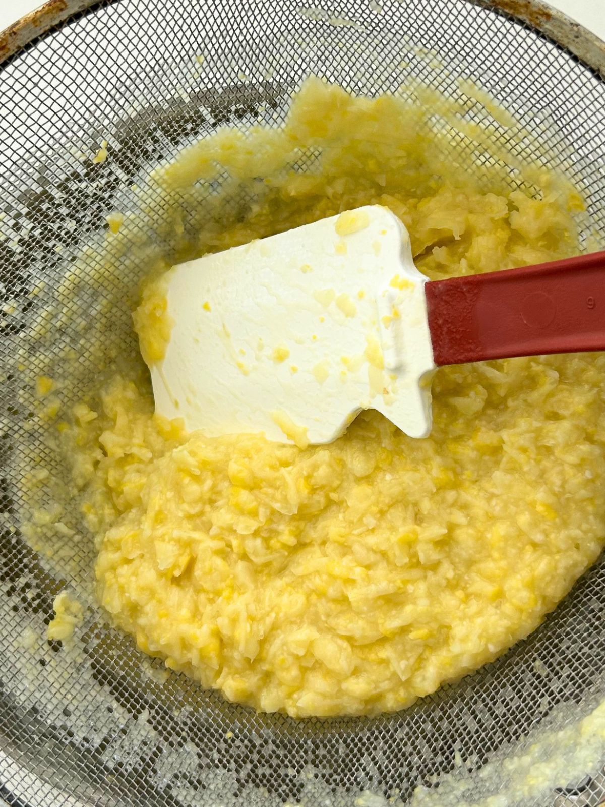 A spatula is pushing down on fiberous vegetables in the strainer leaving behind smooth corn soup in a pot below.