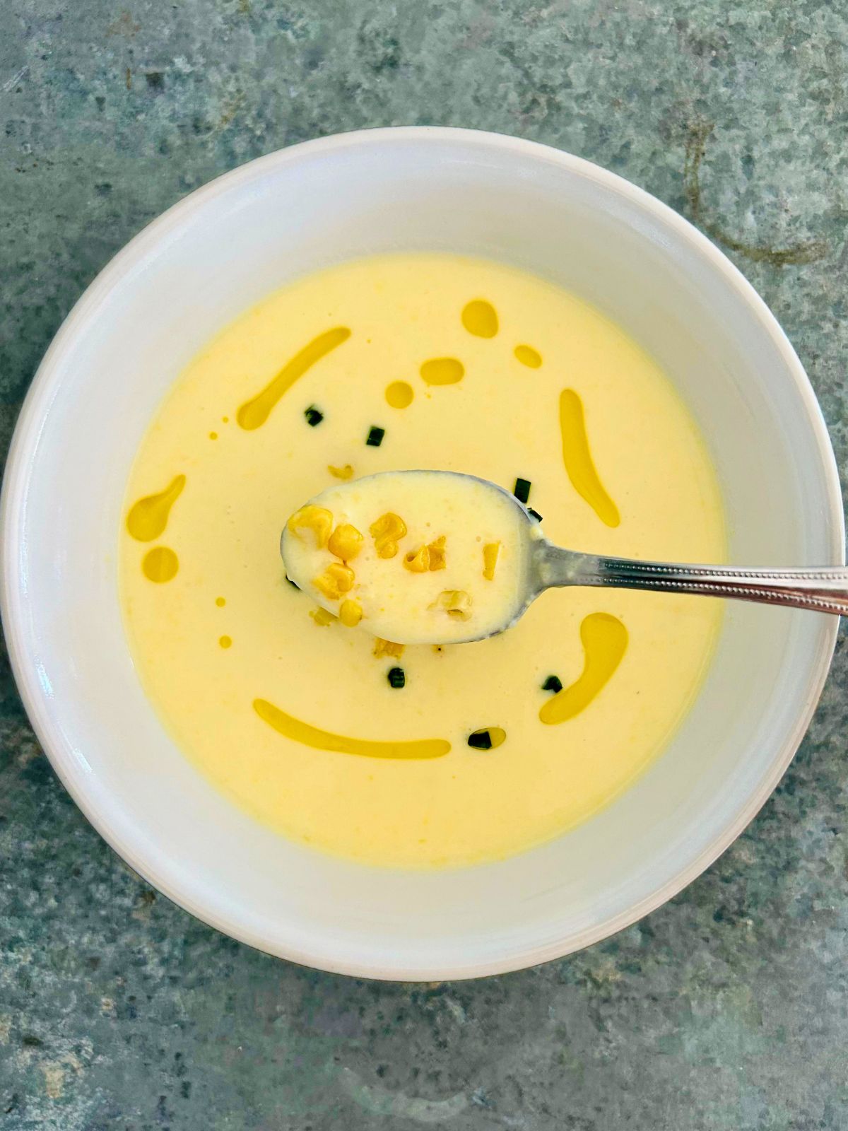 A white bowl with a spoonful of creamy corn soup and a garnish of corn kernels, chives, and olive oil.