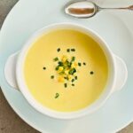 A white bowl of creamy corn soup, chives, basil, and corn kernels next to a silver tablespoon sitting on a white plate.
