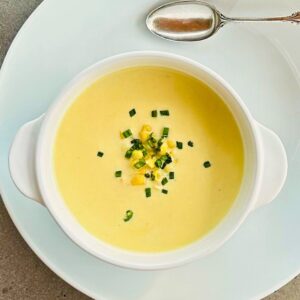 A white bowl of creamy corn soup, chives, basil, and corn kernels next to a silver tablespoon sitting on a white plate.
