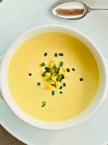 A white bowl of creamy corn soup, chives, basil, and corn kernels next to a silver tablespoon sitting on a white plate.