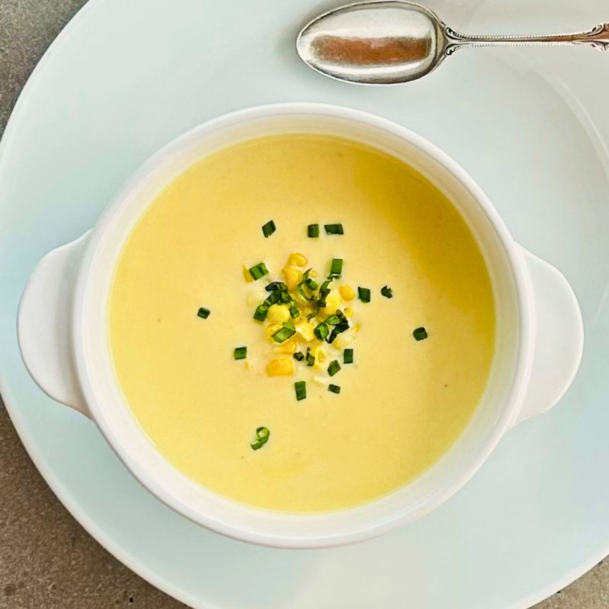A white bowl of creamy corn soup, chives, basil, and corn kernels next to a silver tablespoon sitting on a white plate.