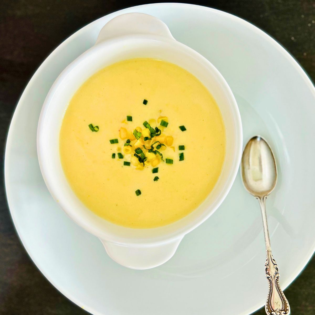 A large white plate holds a spoon and white bowl of creamy corn soup.