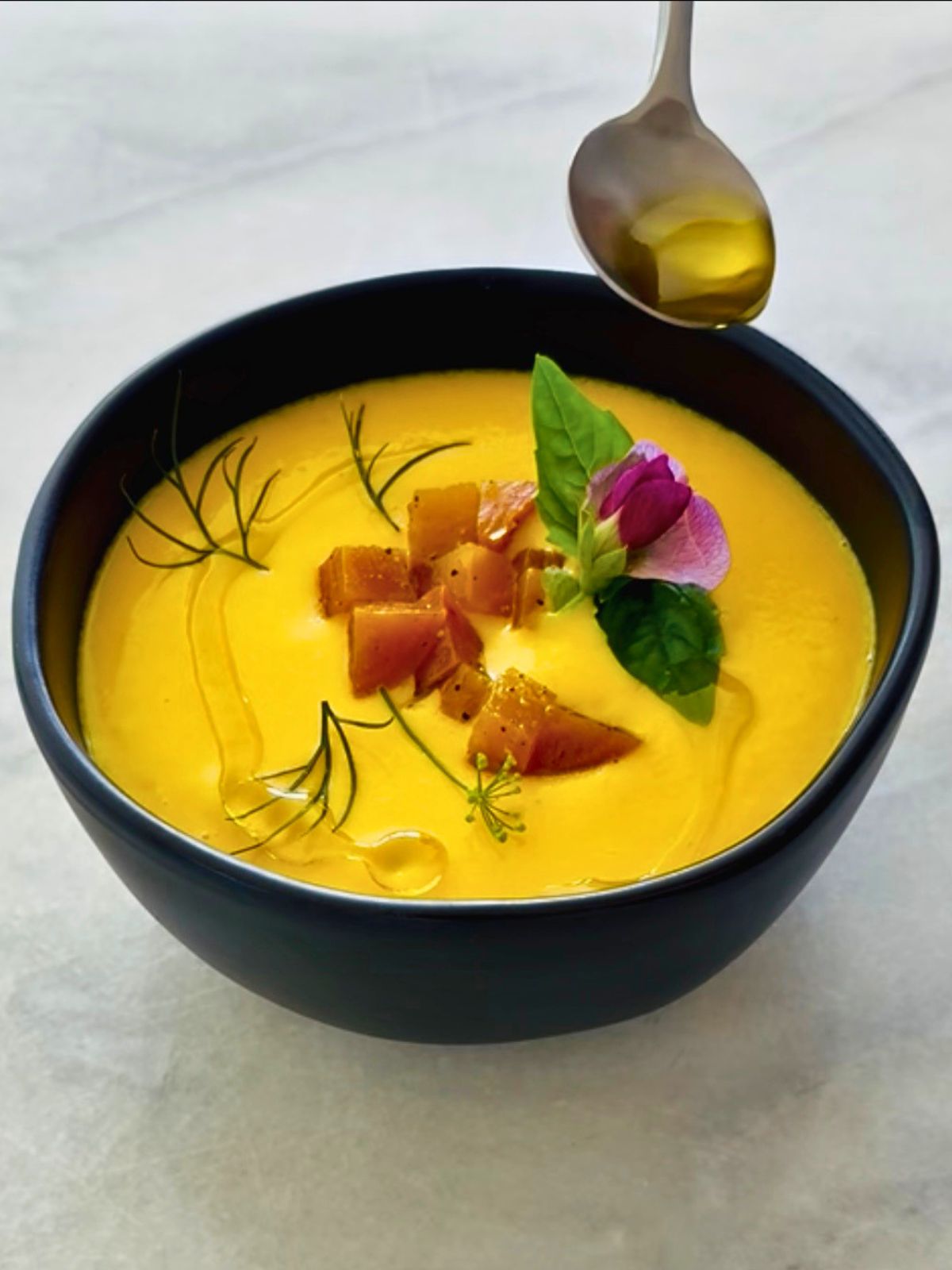 A bowl of golden beet soup garnished with herbs, edible flowers, and olive oil drizzle.
