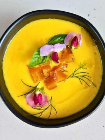 Chilled golden beet soup with buttermilk topped with diced beets, herbs and pea blossoms in a black bowl.