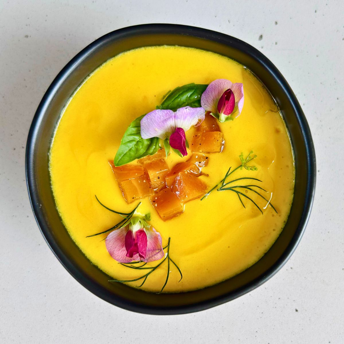 Chilled golden beet soup with buttermilk topped with diced beets, herbs and pea blossoms in a black bowl.