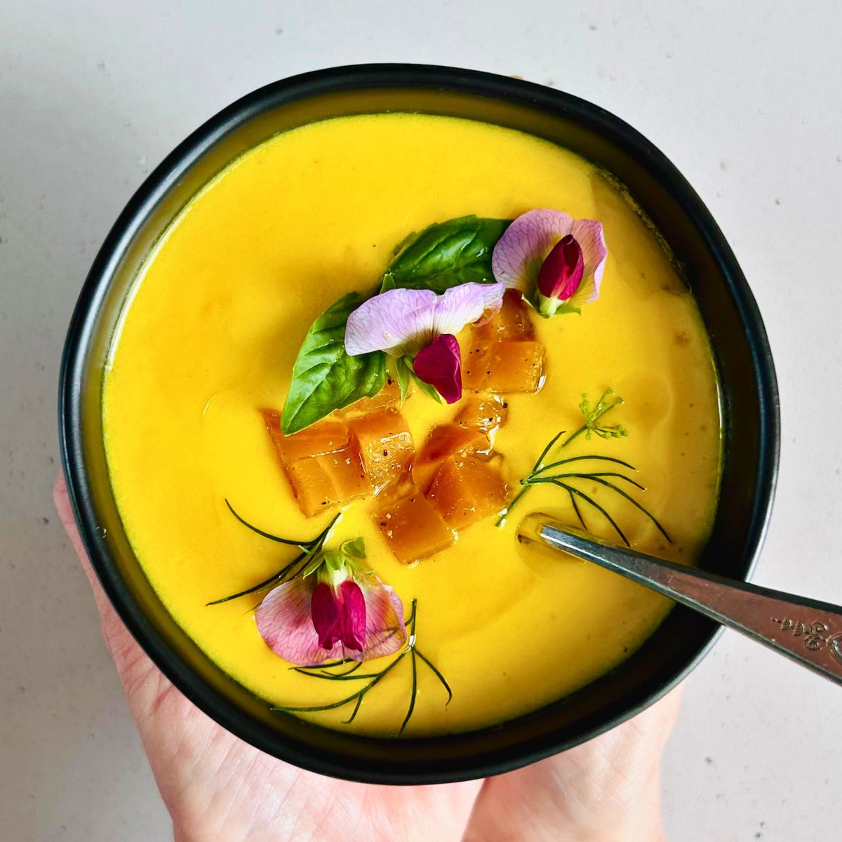 Golden beet soup with a spoon in a black bowl topped with diced beets, dill, basil, and garden pea blossoms.