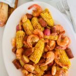 A large white platter holds oven baked shrimp boil next to French bread and forks.