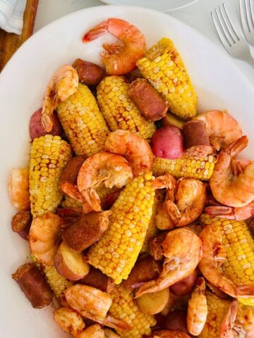 A large white platter holds oven baked shrimp boil next to French bread and forks.