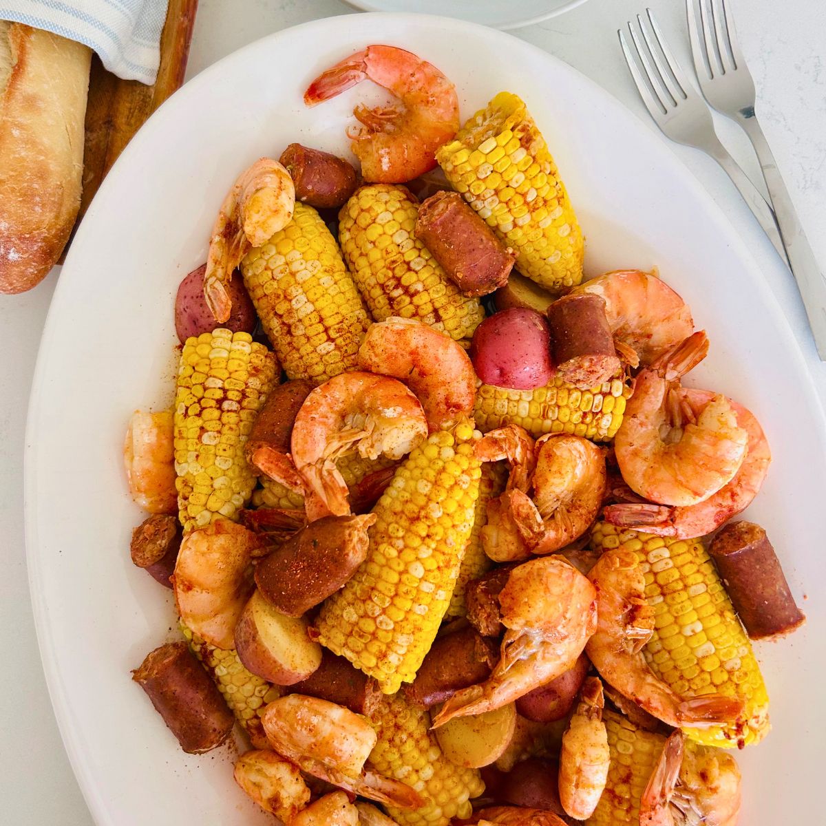 A large white platter holds oven baked shrimp boil next to French bread and forks.