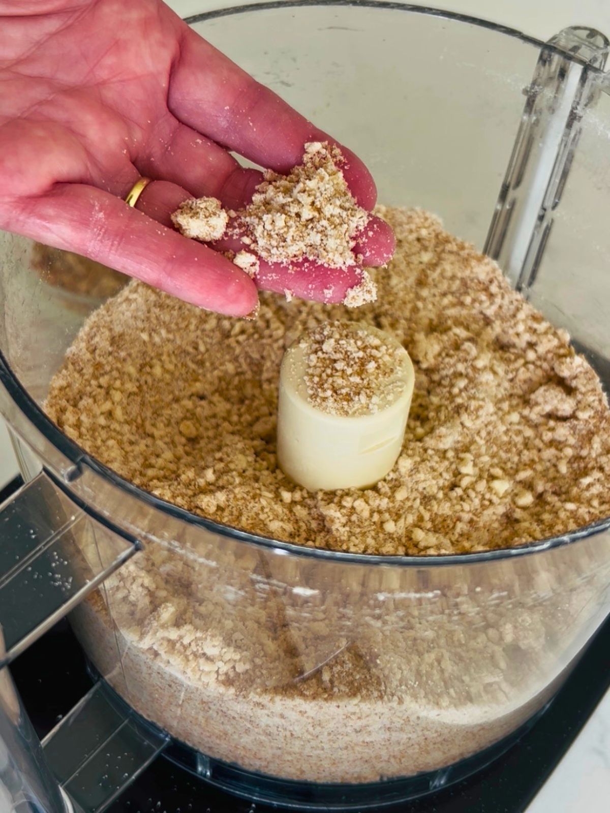 Hand testing the crumbly pecan topping texture over a food processor bowl.