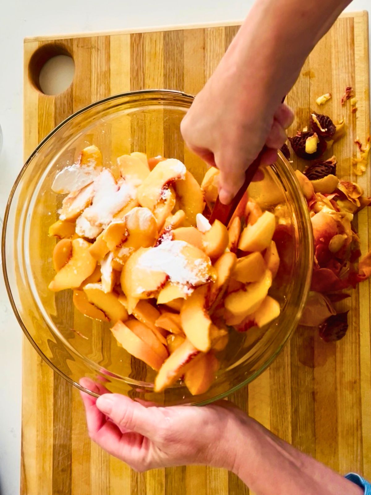 A spatula is tossing sliced fresh peaches with sugar and cornstarch in a large glass bowl with peach skins discarded to the side.