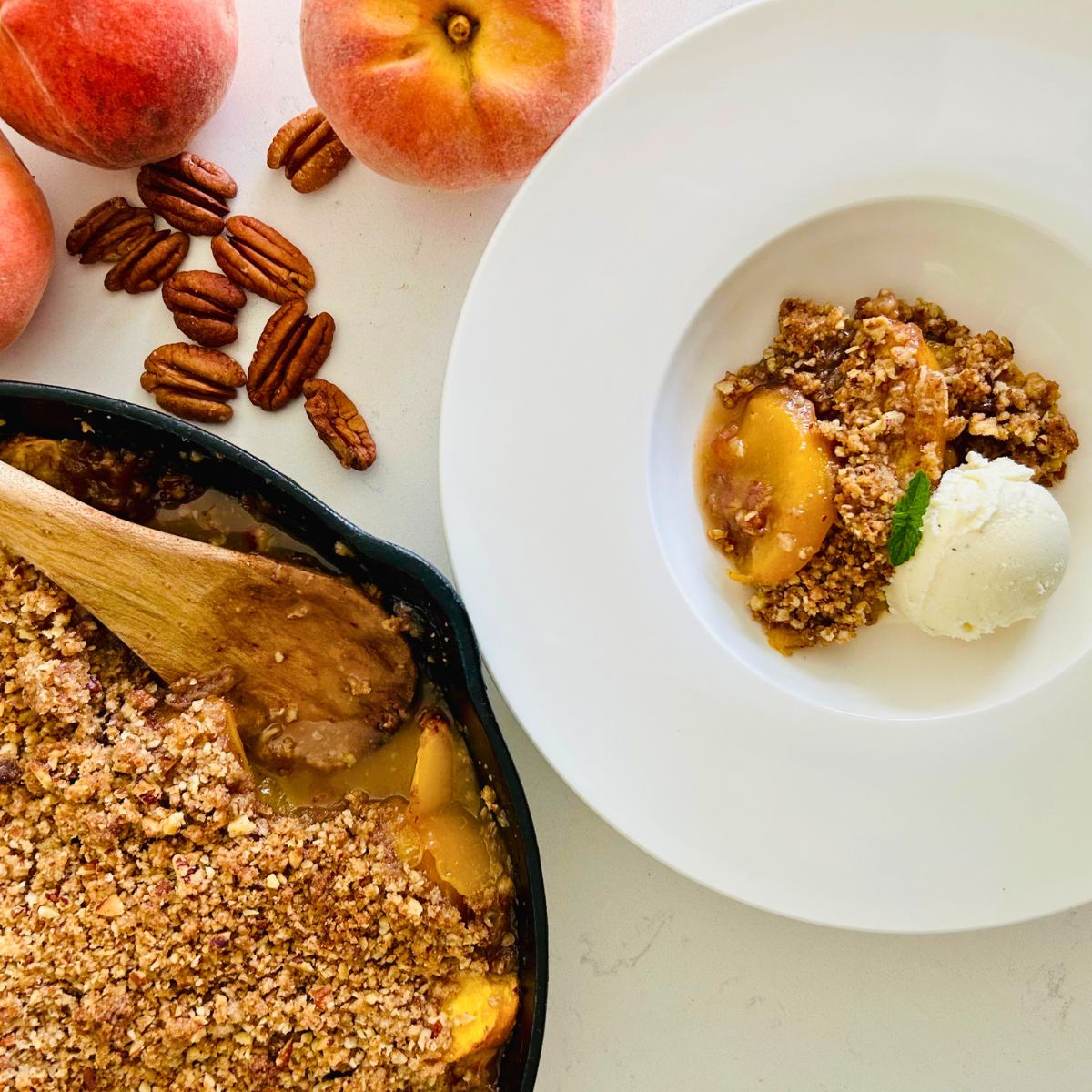 Peach crisp with pecans served in a white bowl with a scoop of vanilla ice cream, alongside the cast iron skillet with a wooden spoon, whole fresh peaches, and loose pecans.