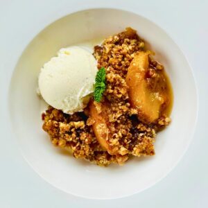 Peach crisp with pecans in a white bowl, topped with a scoop of vanilla ice cream and a fresh mint sprig, showing golden caramelized peach slices and crunchy pecan topping.