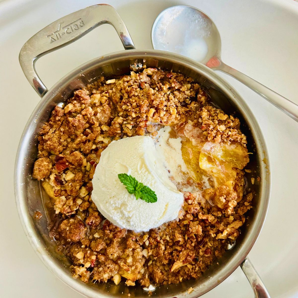 Peach crisp with pecans in an individual sized stainless steel All-Clad sauté pan with a scoop of melting vanilla ice cream, fresh mint and a spoon resting alongside.