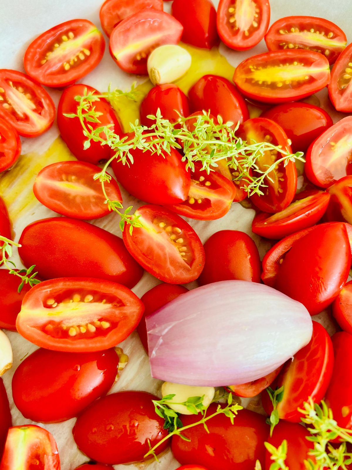 Halved Juliet cherry tomatoes with shallot, garlic, and thyme on a parchment-lined pan ready to roast.