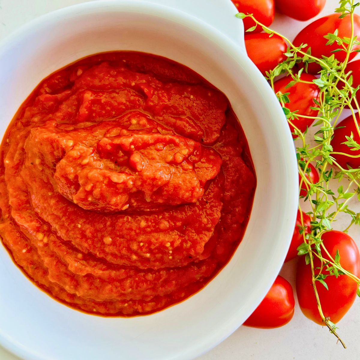 Tomato paste recipe in a white bowl surrounded by fresh Juliet tomatoes and sprigs of thyme on a white surface.