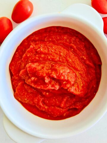 Homemade roasted tomato paste in a white bowl surrounded by fresh Juliet tomatoes on a white marble surface.