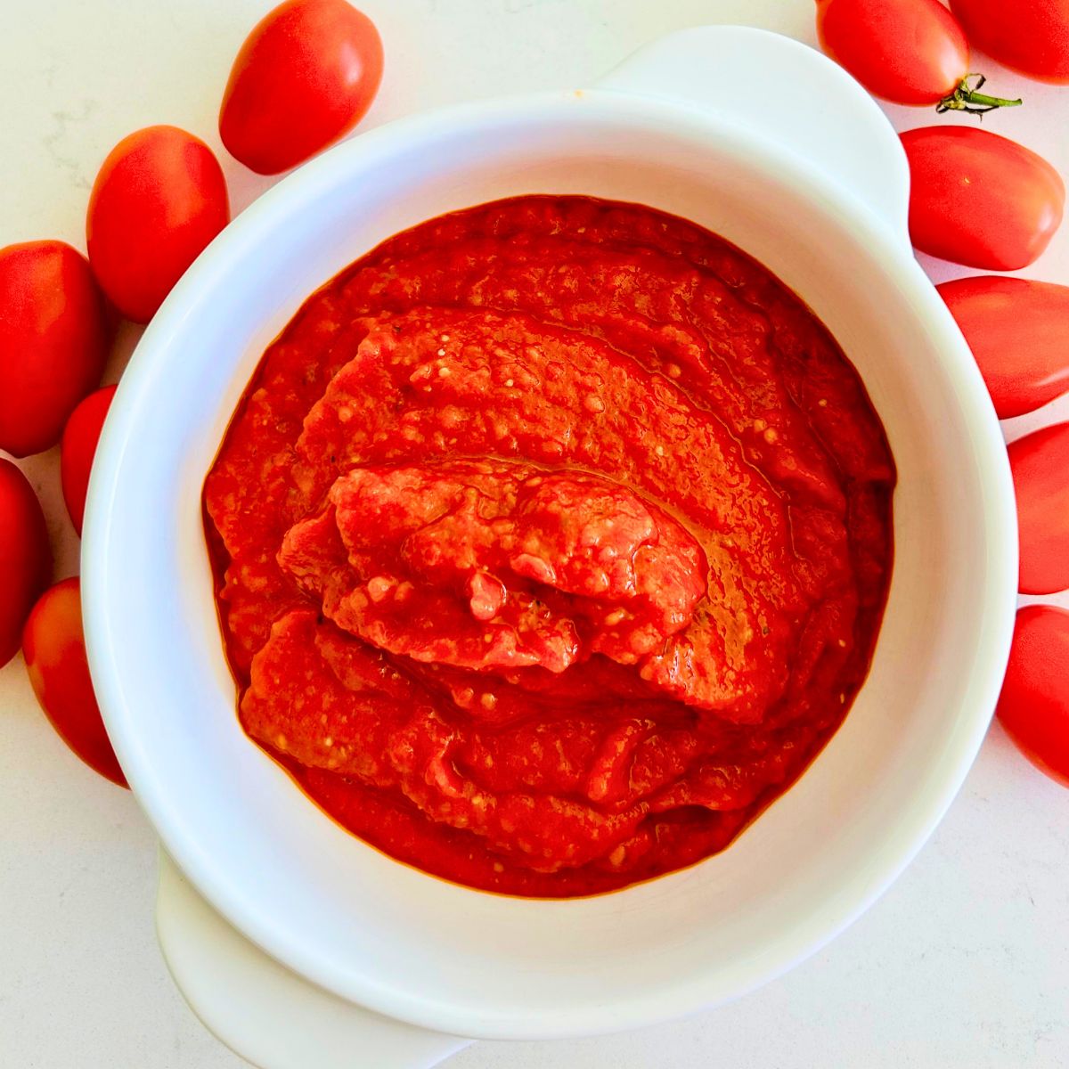 Homemade roasted tomato paste in a white bowl surrounded by fresh Juliet tomatoes on a white marble surface.