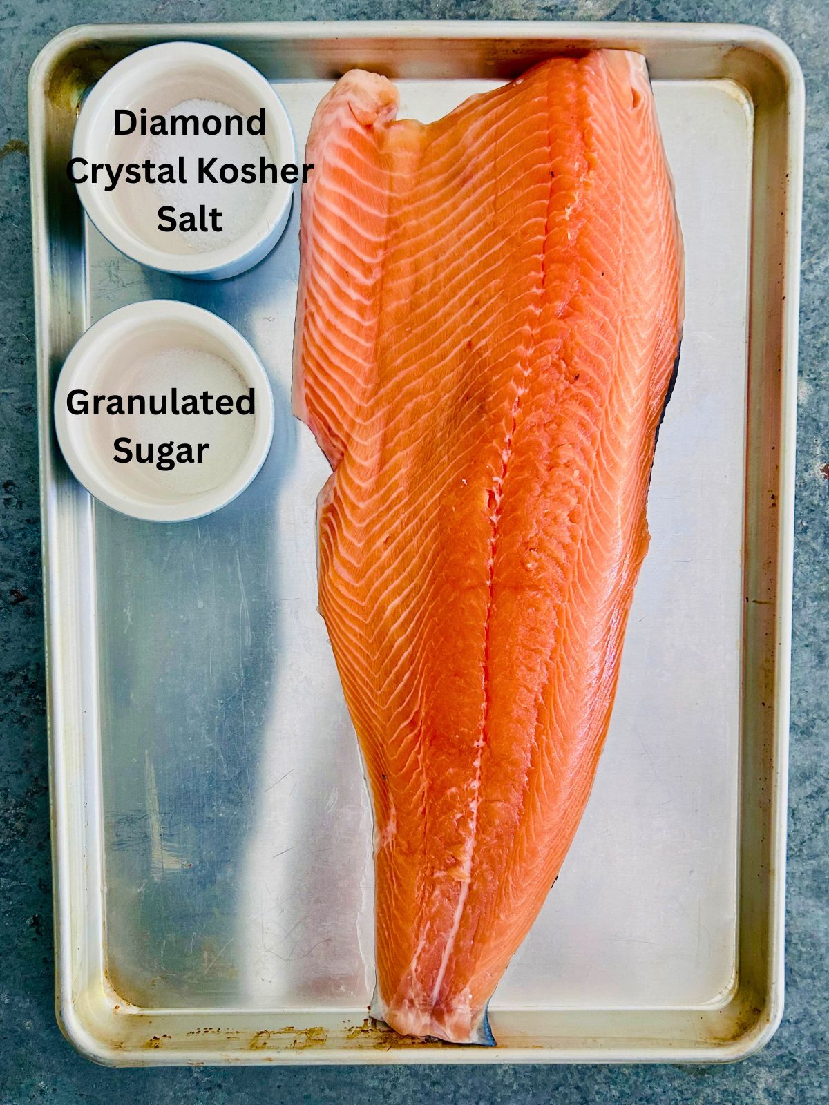 A side of uncooked salmon and bowls of kosher salt and sugar on a rimmed baking sheet.