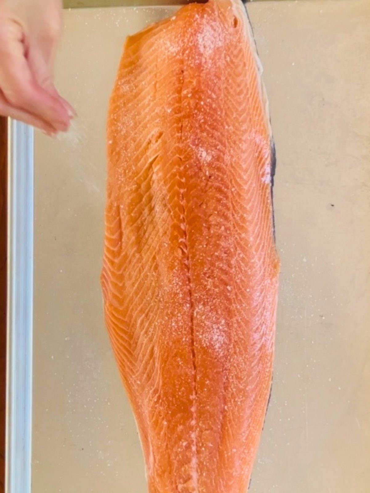 Kosher salt and sugar are sprinkled over a side of salmon on a rimmed baking sheet.