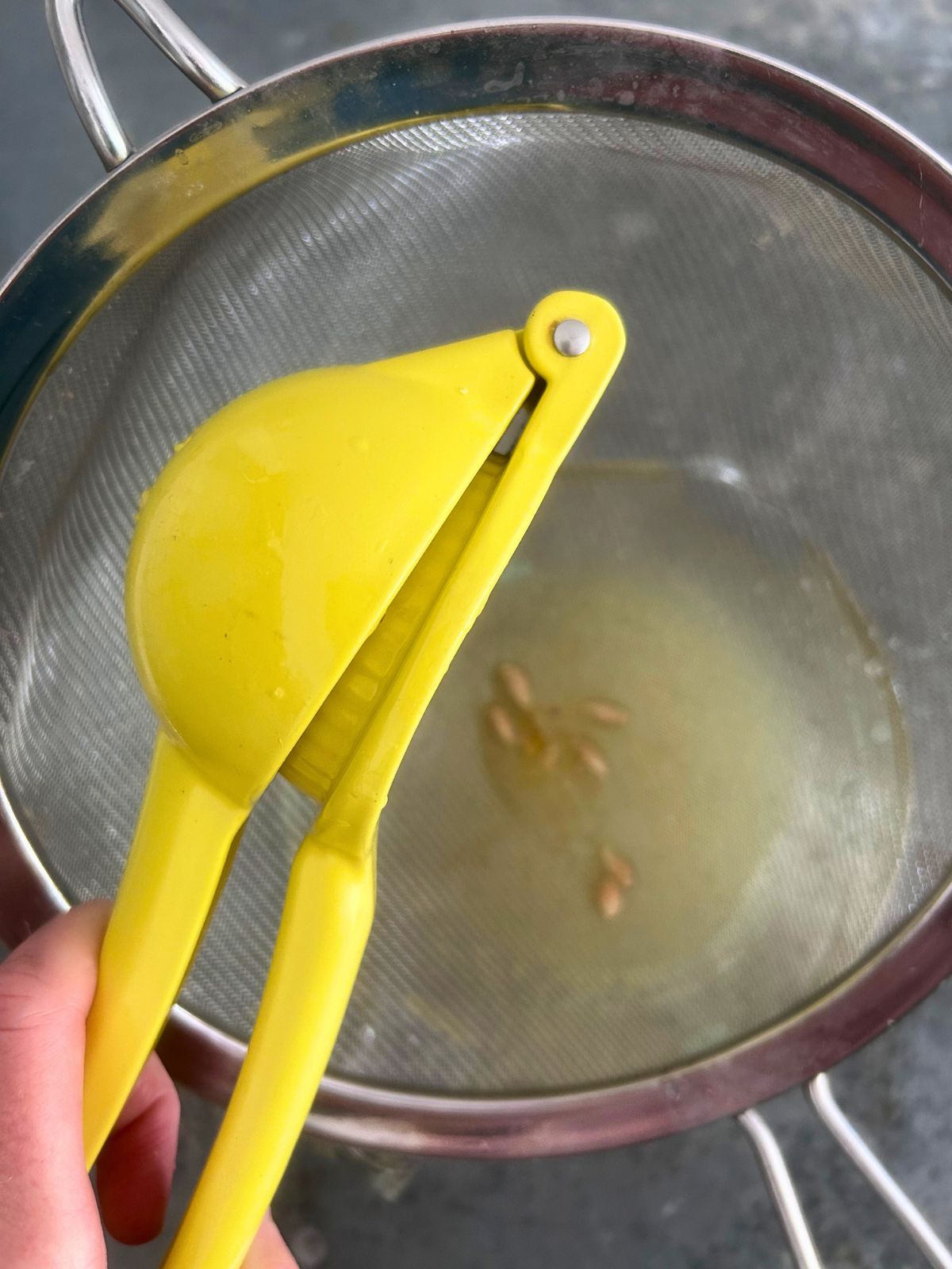 Juicing a lemon with a handheld citrus squeezer over a wire mesh strainer to remove the lemon seeds.