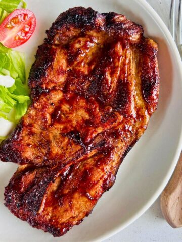 BBQ pork steak on a white plate next to salad and silverware.