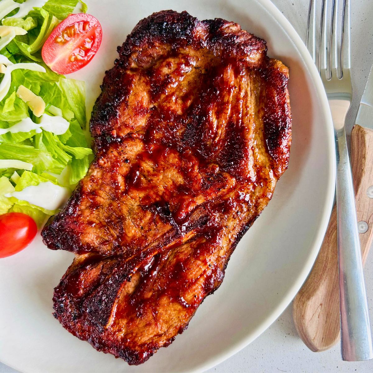 BBQ pork steak on a white plate next to salad and silverware.