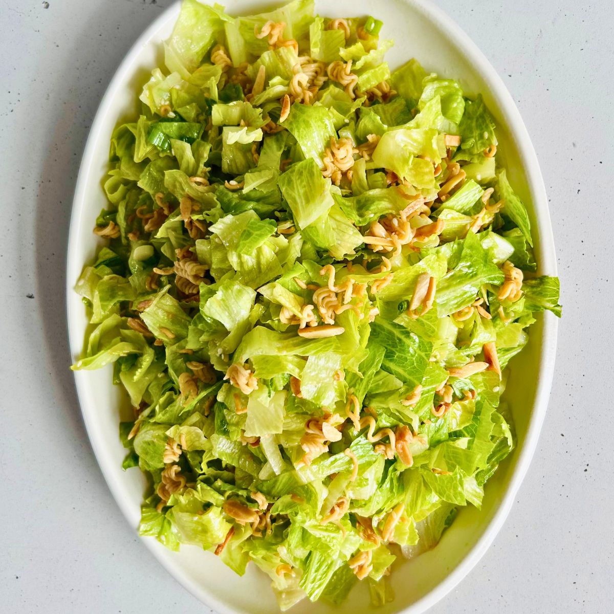 A white oval platter of crunchy romaine salad with ramen noodles, slivered almonds, and Asian sesame dressing.