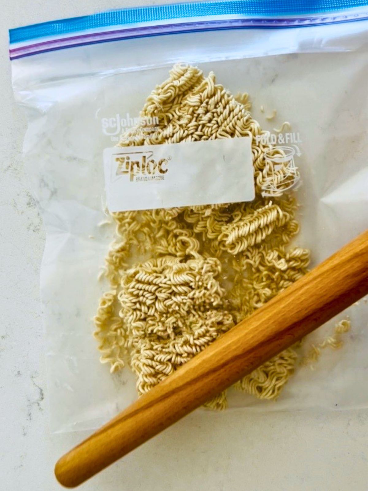 A rolling pin is crushing ramen noodles inside a Ziplock bag.