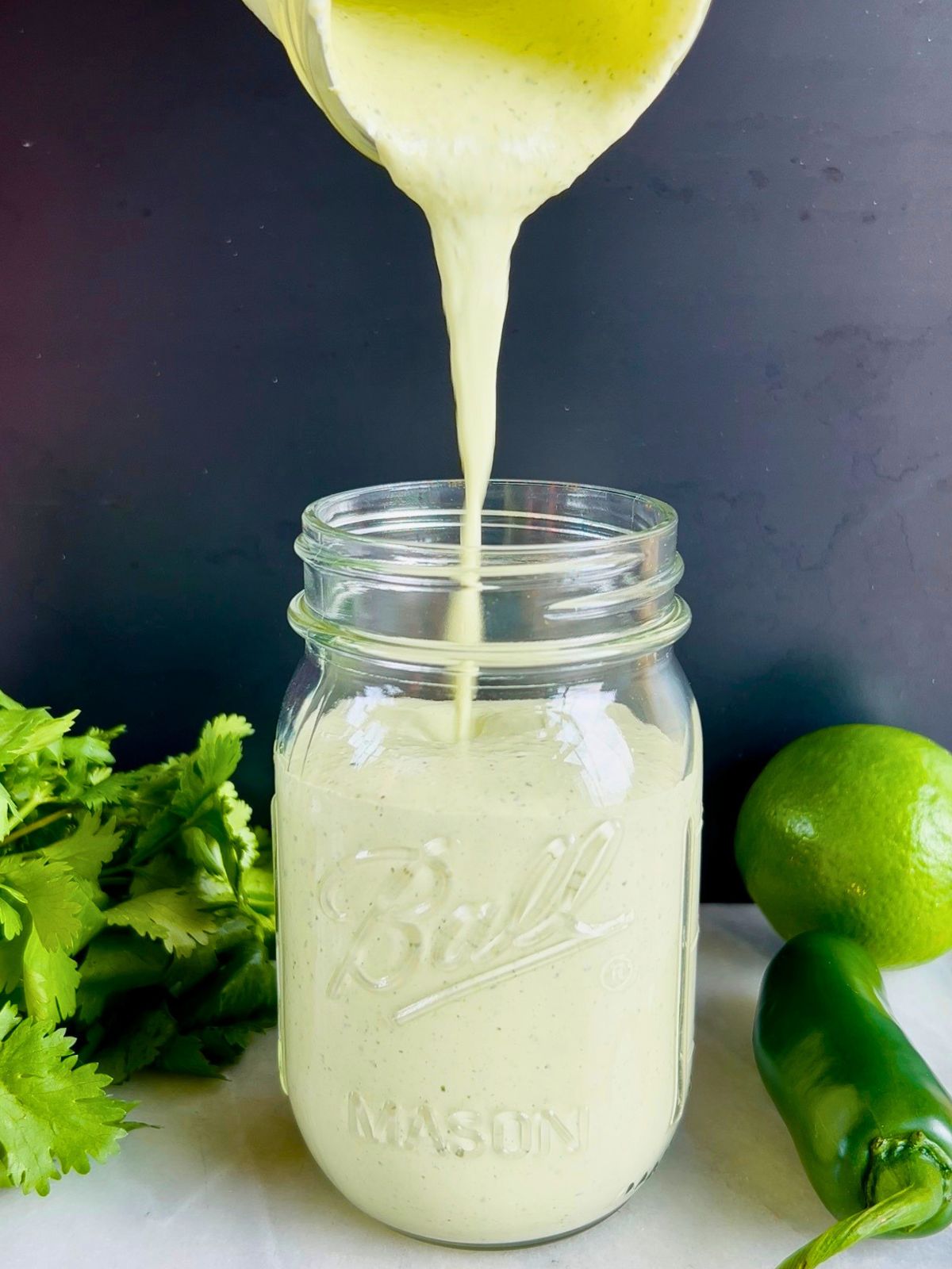 Jalapeño ranch is poured into a mason jar next to cilantro, lime, and a jalapeño pepper.