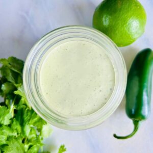 A mason jar of jalapeño ranch dressing next to a bunch of cilantro, lime, and a jalapeño pepper.