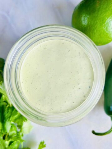 A mason jar of jalapeño ranch dressing next to a bunch of cilantro, lime, and a jalapeño pepper.