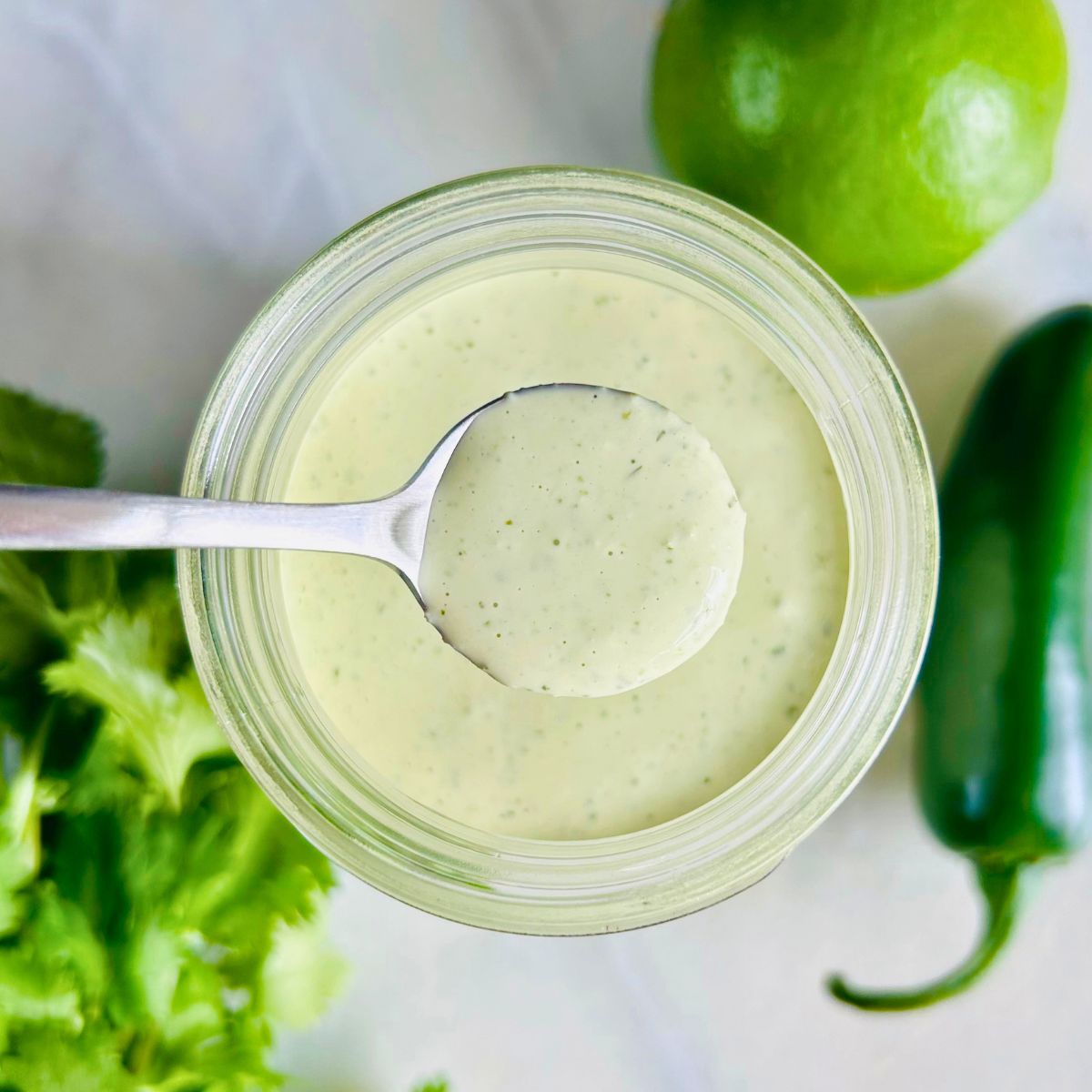 A spoonful of jalapeño ranch held above a mason jar of the dressing along with jalapeño pepper, cilantro, and a lime.