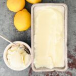 Lemon ice cream in a loaf pan next to lemons and a scoop in a small bowl with a spoon inside.