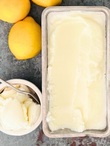 Lemon ice cream in a loaf pan next to lemons and a scoop in a small bowl with a spoon inside.