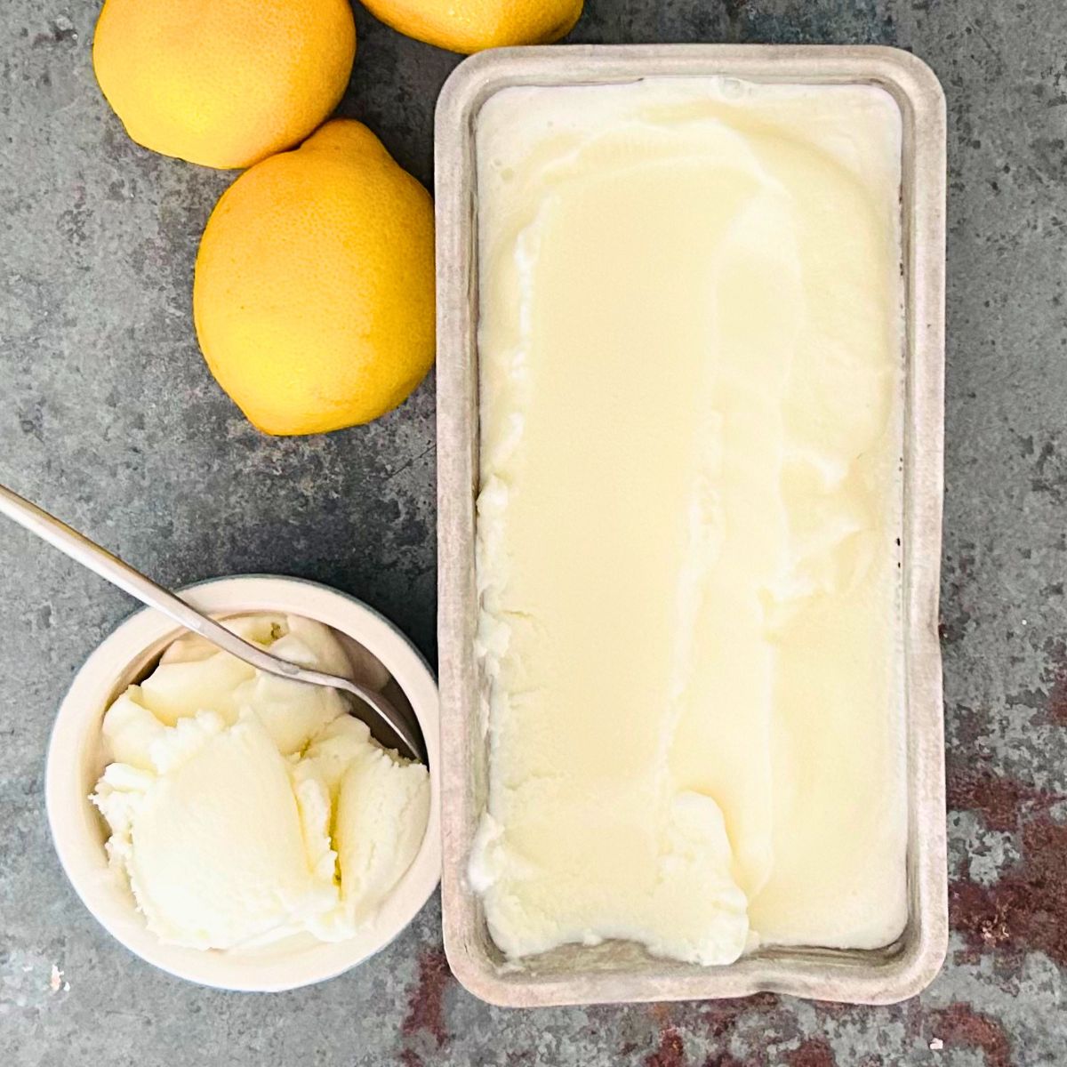 Lemon ice cream in a loaf pan next to lemons and a scoop in a small bowl with a spoon inside.